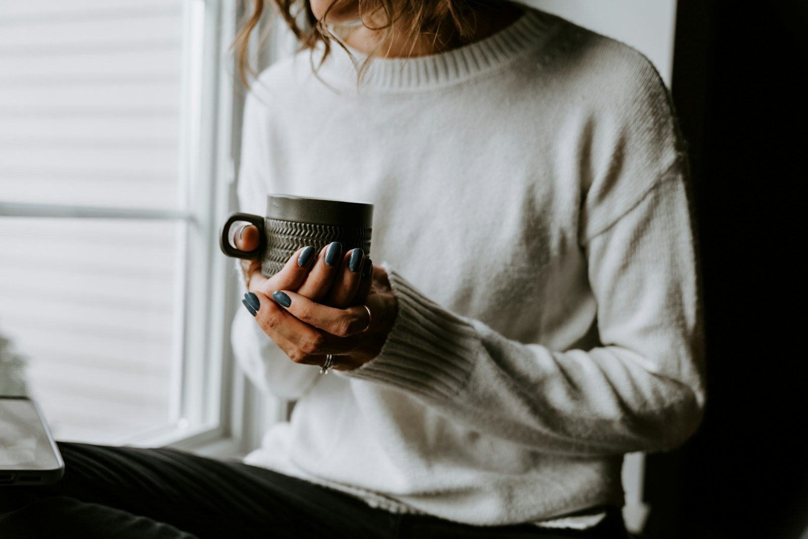 窓辺で白いニットを着た人がマグカップのコーヒーを両手で持ちくつろぐ様子