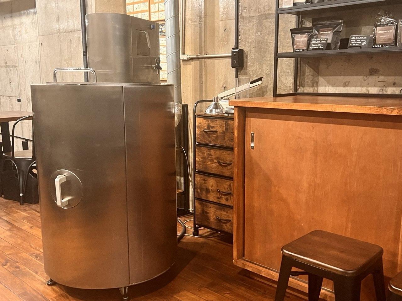 木のカウンターの横に置かれたステンレス製の大型自家焙煎機、コンクリート壁とコーヒー豆の陳列棚が背景に写る店内