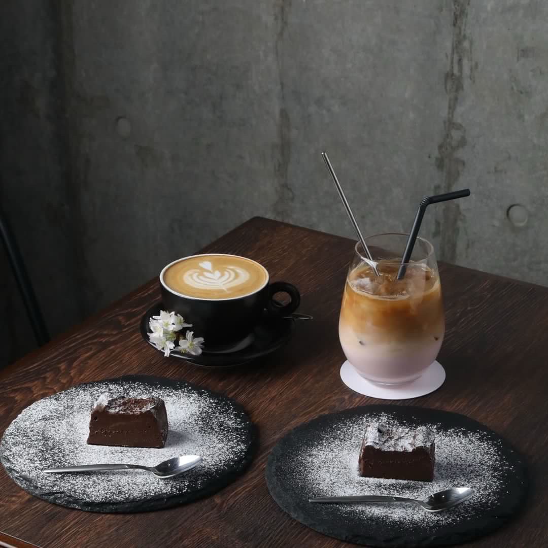 木目のテーブルに並ぶカフェラテとアイスコーヒーそして粉糖をまとったチョコレートケーキ2皿